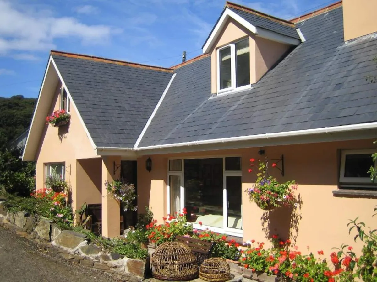 The apricot-rendered front of Kittiwake with its slate roof and dormer window, hanging baskets and geraniums, and willow crab pots beside the bench by the front door